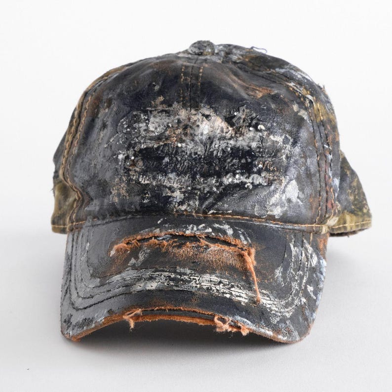 May include: A worn black baseball cap with a curved peak. The cap shows signs of wear and tear, with visible damage and discolouration. The peak has a worn, layered appearance, and the cap's surface has white and silver accents.