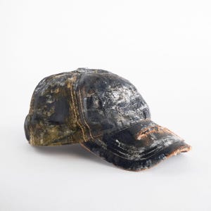 May include: A distressed baseball cap with a worn, textured appearance. The cap has a dark colour scheme with silver and brown accents, and visible stitching. The brim shows signs of wear, adding to its vintage aesthetic.