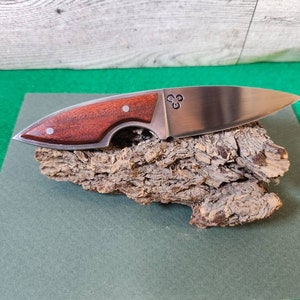 May include: A small, stainless steel knife with a wooden handle. The handle is a dark reddish brown color and has a small, silver circle with three dots in the center. The knife is resting on a piece of bark.