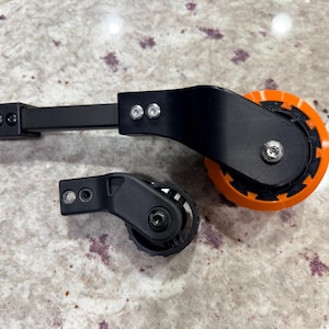 May include: A large black and orange wheel assembly with a metal mounting arm and a smaller black wheel assembly with a matching mounting bracket. Both components feature visible silver screws and are constructed from rigid, durable materials.