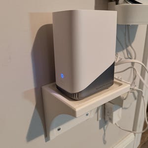 Op de afbeelding: Een witte en grijze rechthoekige router met een blauw lampje, gemonteerd op een witte plank. De router is met een netsnoer en een kleine witte voedingsadapter aan de muur bevestigd.
