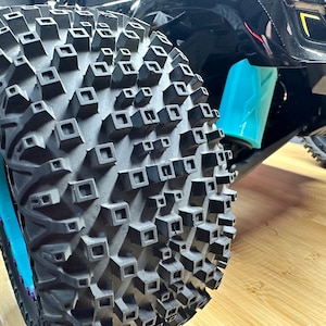 May include: Close-up of a black tyre with a unique tread pattern featuring square-shaped knobs. The tyre is mounted on a vehicle with a blue and white rim.