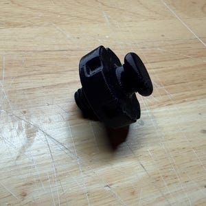 Puede incluir: Un objeto negro impreso en 3D con una base roscada y un botón en la parte superior. El objeto parece ser un tapón de botella o un artículo similar. El tapón está hecho de un material negro mate sólido.