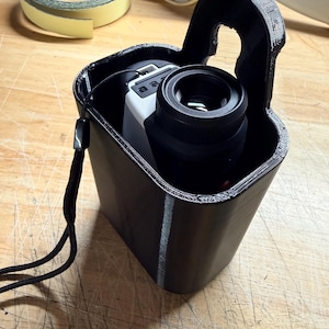 May include: Black 3D-printed carrying case with a handle, holding a white and black device with a lens. A black lanyard is attached. The case is on a wooden surface.