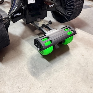 May include: A black and green motorized golf cart with the text "NAV" and "MGI". The cart has a black frame with green wheels and black covers on the wheels. The cart is on a concrete surface.