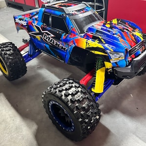 Może przedstawiać: Niebiesko-żółto-czerwony zdalnie sterowany samochód Traxxas X-Maxx. Samochód ma duże, klockowate opony z niebieskimi felgami i żółtymi akcentami. Nadwozie ma kolorowy wzór graficzny. Zawieszenie ma żółte i zielone elementy.