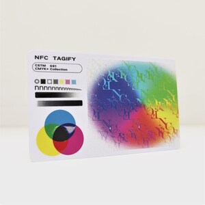 May include: A white card with the text "NFC TAGIFY" and "CSTM 031 CMYK+ Collection" printed on it. The card also features a colour chart with a black circle, a black square, a black and white chequered pattern, a yellow square, a blue square, and a black and white gradient bar. The card also features a colourful design with the text "NFC" repeated in a circular pattern.