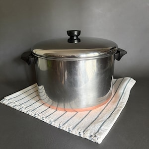 Op de afbeelding: Een roestvrijstalen soeppan met een gepolijste afwerking, voorzien van een koperkleurige bodem, twee zwarte zijhandgrepen en een bijpassend deksel met een zwarte knop. De pan heeft een cilindrische vorm en een glad, reflecterend buitenoppervlak.