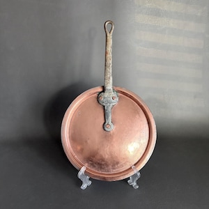 May include: A copper-colored, round pan lid with a long, riveted handle. The lid is displayed on a clear stand against a dark background. The handle is made of a darker metal, and the lid has a shiny, reflective surface.