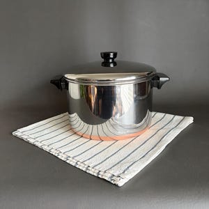 Puede incluir: Una olla de acero inoxidable con tapa y asas negras, colocada sobre un paño a rayas blancas y azules. La olla tiene un fondo cobrizo. El fondo es gris liso.