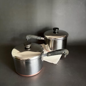 May include: Two stainless steel saucepans with lids. One saucepan has a copper-colored base. Both have black handles and lids with black knobs. The pans are on a dark surface.