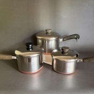 May include: A set of three stainless steel saucepans with copper-colored bases and matching lids. Each pot features a dark, textured handle and a central knob on the lid. The pots vary in size, with one positioned behind the other two in the front.