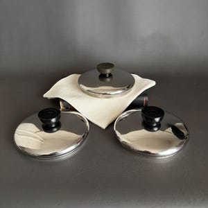 May include: Three stainless steel pot lids with black knobs. One lid is on a white cloth, and the other two are on a gray surface. The lids have a shiny, reflective surface.