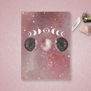 Op de afbeelding: Een roze en wit notitieboek met een galaxy-ontwerp en een maan- en sterillustratie. Het notitieboek heeft een wit oog in het midden van de ster.