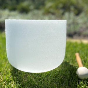 Puede incluir: Un cuenco de cristal de canto esmerilado y transl&uacute;cido con base redondeada sobre hierba verde. Un mazo de madera con cabeza blanca descansa cerca. El cuenco est&aacute; dise&ntilde;ado para la curaci&oacute;n por sonido y la meditaci&oacute;n.