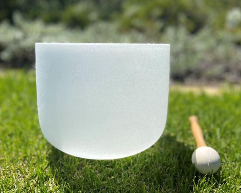 Puede incluir: Un cuenco de cristal de canto esmerilado y transl&uacute;cido con base redondeada sobre hierba verde. Un mazo de madera con cabeza blanca descansa cerca. El cuenco est&aacute; dise&ntilde;ado para la curaci&oacute;n por sonido y la meditaci&oacute;n.