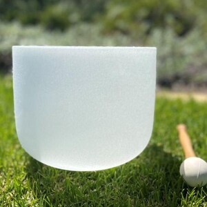 Puede incluir: Un cuenco de cristal blanco esmerilado con base redondeada sobre hierba verde. Un mazo de madera con cabeza blanca est&aacute; cerca. El cuenco se utiliza para la curaci&oacute;n por sonido y la meditaci&oacute;n.