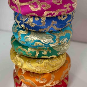 以下が含まれることがあります： 8つのカラフルな瞑想クッションの積み重ね。それぞれ異なる刺繍のデザインが施されています。クッションは虹色のパターンで配置されており、一番上がピンク、一番下が赤になっています。