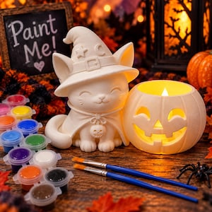 May include: A white ceramic cat figurine wearing a witch hat and cape with a small skull charm, next to a white ceramic jack-o'-lantern candle holder with a glowing light, paint pots, and two paintbrushes. A sign reads Paint Me!