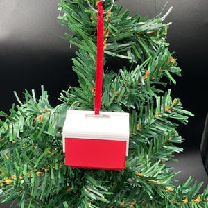Puede incluir: Un adorno navideño en miniatura con forma de nevera roja y blanca. El adorno cuelga de un lazo de metal plateado con una cinta roja. El adorno está colocado sobre un fondo de árbol de Navidad verde.