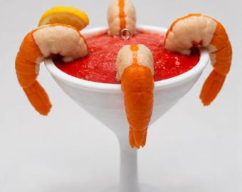 May include: A white martini glass filled with a textured red cocktail sauce, garnished with four orange and beige shrimp and a yellow lemon slice. A small silver metal eyelet is attached to the top center for hanging as an ornament.