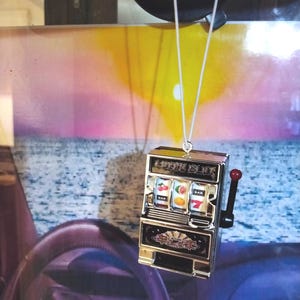 May include: A silver-colored slot machine pendant with a red handle and white string. The slot machine displays fruit symbols and the word "BAR". The background features a sunset over the ocean.