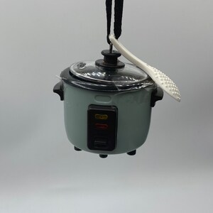 Może przedstawiać: Miniaturowy, jasnozielony garnek do gotowania ryżu z przezroczystą pokrywką i czarnymi akcentami. Biała łopatka do ryżu spoczywa z boku. Małe urządzenie wisi na czarnym sznurku na białym tle. Posiada czerwone i żółte kontrolki.