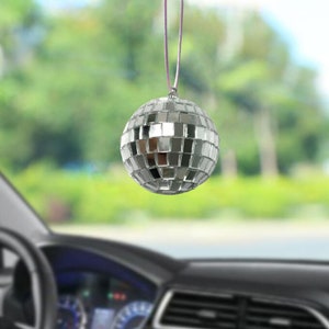 May include: A silver disco ball hanging from a purple string, reflecting light. The spherical ornament is made of small, mirrored squares. The background is a blurred view of a car interior and a green, leafy outdoor scene.