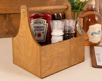 May include: Wooden caddy with a handle, holding a bottle of ketchup, two salt and pepper shakers, and a bottle of wine. The caddy is on a wooden table with a white tablecloth.