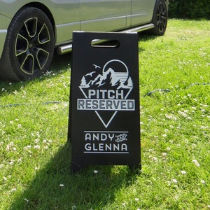 May include: A silver campervan with a red pop-up roof is parked on grass. A black sign with white text reading "PITCH RESERVED" and the names "ANDY and GLENNA" stands in front. The van is connected to a blue power cable.