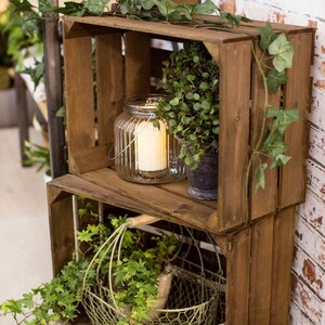 May include: Two stacked wooden crates with a wire basket and a glass candle lantern on the top shelf. The crates are decorated with green ivy vines.