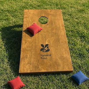 May include: A rectangular wooden cornhole board with a circular hole at the top, featuring a black National Trust logo and text printed in the centre. One red bean bag rests on the board, while another red bag and a blue bag lie nearby.