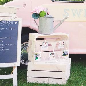 May include: A white wooden crate with a galvanized metal watering can and a string of photos hanging from clothespins. The crate is sitting on a green grassy surface. The text "SWE" is visible on a pink surface in the background.