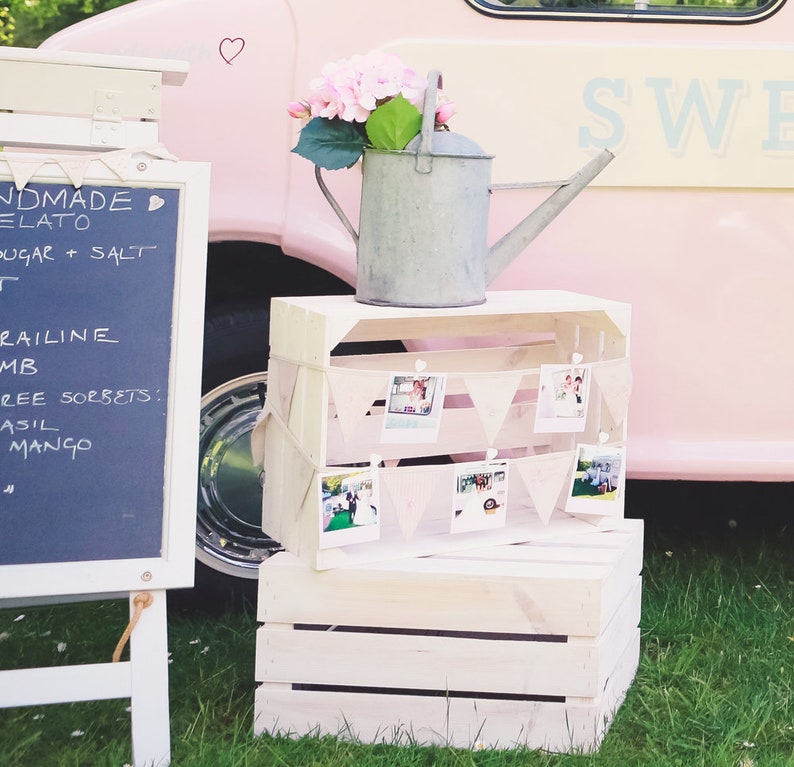 May include: A white wooden crate with a galvanized metal watering can and a string of photos hanging from clothespins. The crate is sitting on a green grassy surface. The text "SWE" is visible on a pink surface in the background.