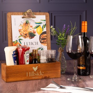 May include: A wooden condiment holder with the words "Hillside Bar & Inn" engraved on the front. The holder contains a bottle of ketchup, a bottle of mayonnaise, a bottle of soy sauce, a salt shaker, and a pepper shaker. A wooden clipboard with a menu for lunch is leaning against the holder. A glass of red wine and a bottle of red wine are on the table.