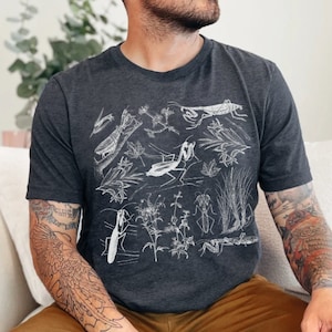 Puede incluir: Camiseta gris oscuro con una ilustración blanca de insectos y plantas. La camiseta de manga corta tiene cuello redondo y está hecha de un material suave. La camiseta se usa con pantalones marrones.