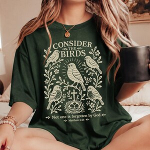 Könnte beinhalten: Dunkelgrünes T-Shirt mit dem Text "Consider the Birds" und einer Grafik von Vögeln und Blättern. Das Shirt enthält auch den Bibelvers Matthäus 6:26. Eine schwarze Tasse wird in der Hand gehalten.