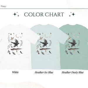 May include: A colour chart shows three t-shirts: white, heather ice blue, and heather dusty blue. Each shirt features a watercolour design of a crow in flight, surrounded by botanical elements. The text "COLOR CHART" is at the top.