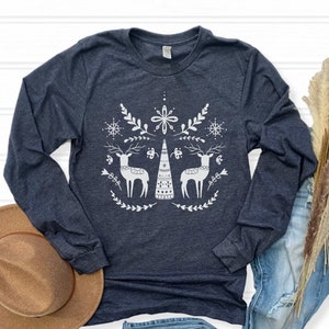 May include: A navy blue long-sleeved t-shirt with a white design of two stylized reindeer flanking a stylized Christmas tree. The design also includes snowflakes and floral elements.