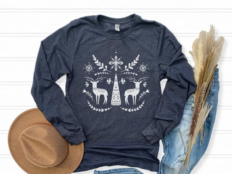 May include: A navy blue long-sleeved t-shirt with a white design of two stylized reindeer flanking a stylized Christmas tree. The design also includes snowflakes and floral elements.
