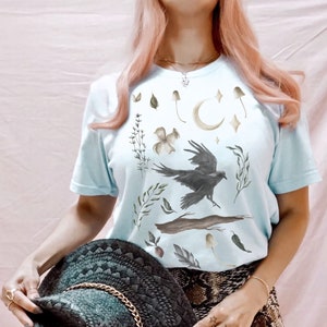 May include: Pale blue t-shirt with a watercolour design of a raven, mushrooms, leaves, and a crescent moon. The shirt has short sleeves and a crew neck. The overall theme is nature and fantasy.