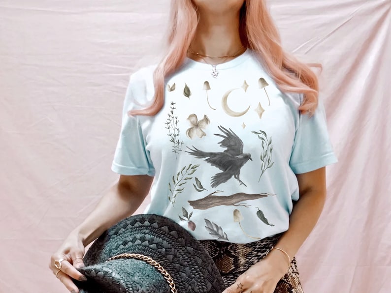 May include: Pale blue t-shirt with a watercolour design of a raven, mushrooms, leaves, and a crescent moon. The shirt has short sleeves and a crew neck. The overall theme is nature and fantasy.