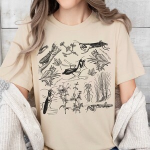 May include: Beige t-shirt featuring a detailed black and white illustration of various insects, including praying mantises, and botanical elements. The design includes insects, leaves, and plants. The shirt is made of a soft material.