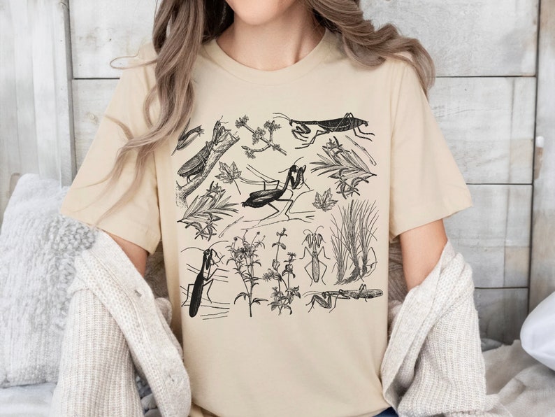 May include: Beige t-shirt featuring a detailed black and white illustration of various insects, including praying mantises, and botanical elements. The design includes insects, leaves, and plants. The shirt is made of a soft material.