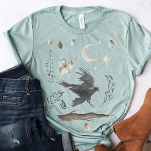 May include: A pale teal t-shirt with a watercolour design of a raven, moon, mushrooms, and floral elements. The shirt is styled with dark wash denim jeans and brown ankle boots, creating a casual, bohemian aesthetic.