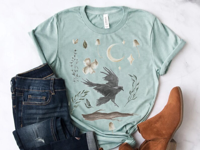 May include: A pale teal t-shirt with a watercolour design of a raven, moon, mushrooms, and floral elements. The shirt is styled with dark wash denim jeans and brown ankle boots, creating a casual, bohemian aesthetic.
