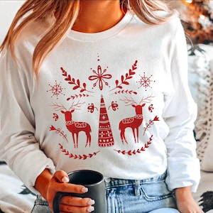 May include: White long-sleeved sweatshirt with a red Christmas design featuring two reindeer, a stylized Christmas tree, snowflakes, and floral elements.