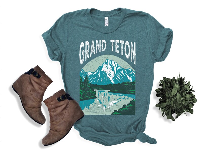May include: Teal t-shirt with a graphic of the Grand Teton mountain range, waterfall, and trees. The text "GRAND TETON" is arched above the mountain scene. Brown wedge ankle boots with buckles are next to the shirt.