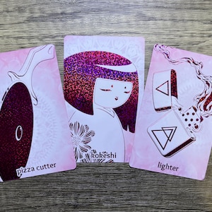 May include: Three pink cards with black line drawings of a pizza cutter, a Japanese doll, and a lighter. The cards have the words "pizza cutter", "Rok&eacute;shi", and "lighter" printed on them.