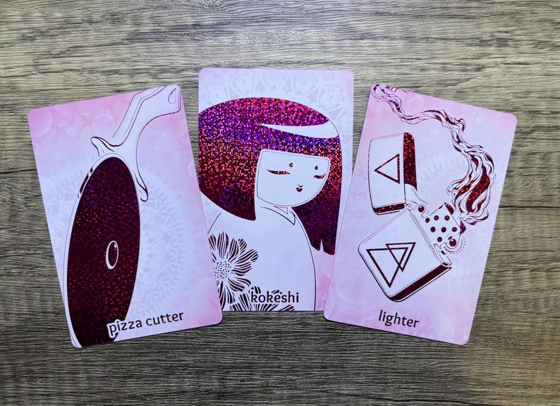 May include: Three pink cards with black line drawings of a pizza cutter, a Japanese doll, and a lighter. The cards have the words "pizza cutter", "Rok&eacute;shi", and "lighter" printed on them.